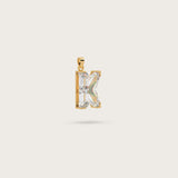 Gold pendant in the shape of the letter "K," adorned with sparkling gemstones, displayed on a light beige background.